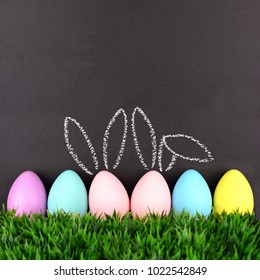 Row of Easter eggs in grass against a chalkboard background. Two with hand drawn Easter Bunny ears.