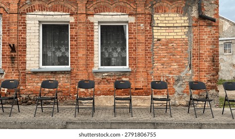 A row of empty black plastic folding chairs lined up on a paved sidewalk against a weathered, rustic red brick building facade featuring old windows with lace curtains and visible structural cracks.