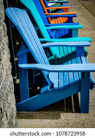 Row Of Empty Colored Adirondack Chairs Against A Gray Brick Wall On A Sidewalk