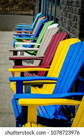 Row Of Empty Colored Adirondack Chairs Against A Gray Brick Wall And Storefront Windows On A Sidewalk