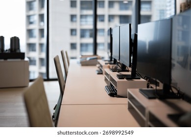 A row of empty desks with computer monitors and chairs in a modern, sunlit office with a city view.