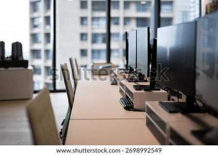 A row of empty desks with computer monitors and chairs in a modern, sunlit office with a city view.