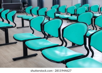 A row of empty turquoise plastic chairs with black metal frames in waiting area, arranged in neat lines on light wooden floor, creating calm and orderly atmosphere