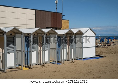A row of functional white changing cabins sits on a sandy beach, offering visitors privacy while enjoying the seaside, with sun loungers and beachgoers visible in the background.