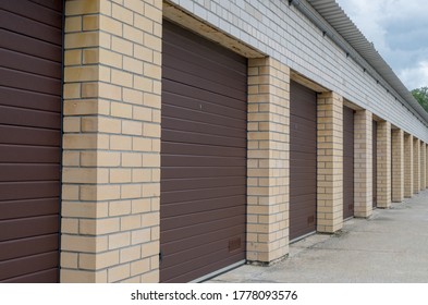 Row of garage boxes under one roof with walls of white brick with brown accordion folding doors, with drainpipe along entire length goes into perspective on background of concrete floor and cloudy sky