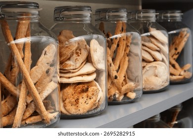 Row of glass jars filled with assorted breadsticks and crackers on a shelf, showcasing rustic baked goods in clear storage with shallow depth of field.