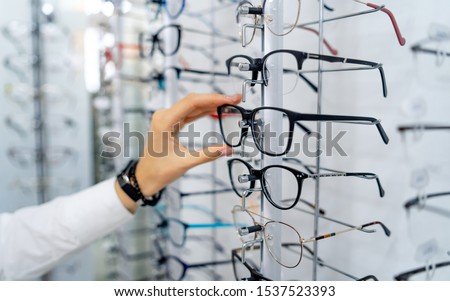 Row of glasses at an opticians. Eyeglasses shop. Stand with glasses in the store of optics. Woman's hand chooses spectacles. Eyesight correction.