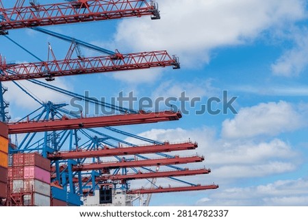 Row of large red container gantry cranes against blue sky with clouds at a busy shipping port terminal.