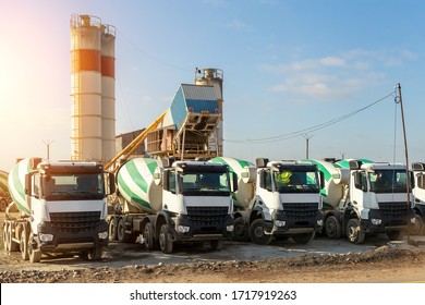 Row Of Many Modern Big Mixer Trucks Parked Against Mobile Temporary Concrete Plant Factory At New Asphalt Road Construction Site Morning Day. Heavy Machniery And Industrial Facilities Background