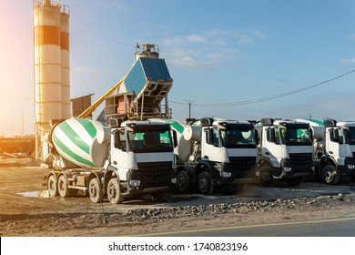 Row Of Many Modern Big Mixer Trucks Parked Against Mobile Temporary Concrete Plant Factory At New Asphalt Road Construction Site Morning Day. Heavy Machniery And Industrial Facilities Background.