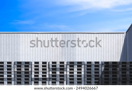 Row of many plastic pallets stacked outside of aluminium warehouse building wall against blue sky background