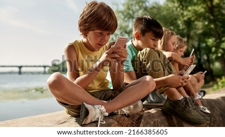 Row of multiethnic children using and browsing mobile phones on log on river coast outdoors. Boys and girl of generation alpha. Gadget addiction. Childhood lifestyle. Sunny summer day