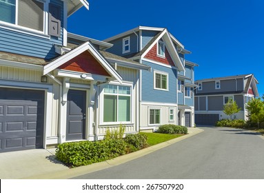 Row Of New Residential Townhouses On A Street On Sunny Day. Family Townhouses With Concrete Driveway And Asphalt Road In Front. British Columbia, Canada.