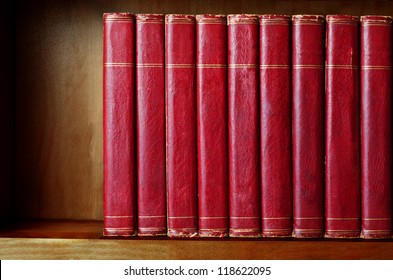 A row of old, battered, encyclopaedias on a shelf, with titles removed.  Red leather effect with gold striped trims.  Shelf has been darkened artificially to give impression of age.