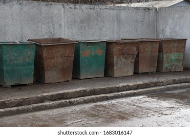 a row of old iron rusty garbage cans stand against a gray concrete wall on asphalt on the street