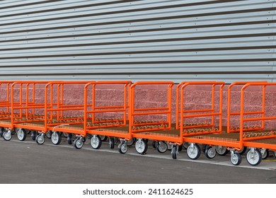 A row of orange color stainless steel heavy-duty trolley carts is stored outside a store with a grey metal exterior wall. The push caddy buggies have four castors on the bottom of the wagon's platform