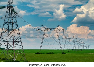 Row Of Power Line Support Pylons On The Green Cultivated Field With Cloudy Sky. Energy Transition Equipment. 