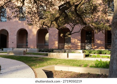 Row of rectangular concrete benches sit among gravel paths and green patches live oak sprawling tree. Arched windows, doorways define red brick building traditional campus architecture, Waco, TX. USA