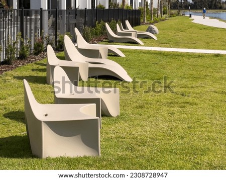 Row of rotationally molded plastic lounge chairs, all the same shade of beige, on green lawn between residential iron fence and lakeside walkway on a sunny morning in southwest Florida