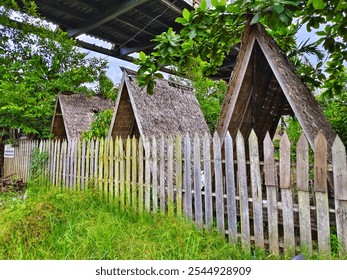 A row of rustic wooden cabins with thatched roofs nestled in lush greenery. A wooden fence lines the foreground, enhancing the rural and serene atmosphere of the setting. - Powered by Shutterstock