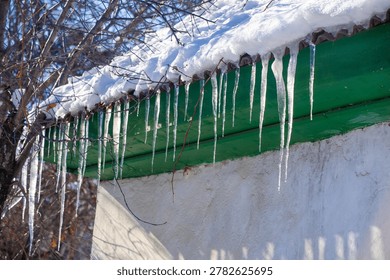 A row of sharp, clear icicles hangs from a snow-covered green roof over a white stucco wall. The winter sunlight casts long shadows and makes the ice glisten.