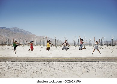 A Row Of Six Young People Leaping In The Air, Arms Outstretched In Wide Open Space In The Desert