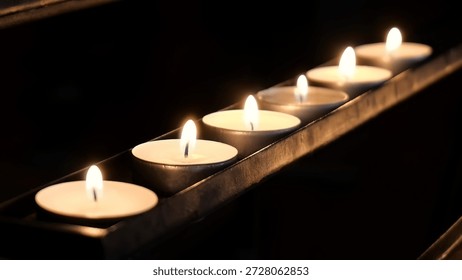 Row of small lit tea light candles in metal holder glowing in dark room, church, warm flame light shallow depth of field and soft bokeh, votive candle prayer meditation memorial atmosphere copy space