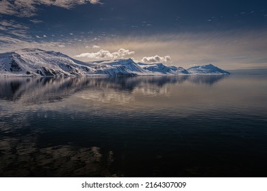 A ROW OF SNOW COVERED MOUNTAINS WITH A REFLECTION IN THE ARCTIC OCEAN AND NUMEROSU CLOUDS NEAR THE ISLAND OF SVALBARD NORWAY IN THE ARCTIC