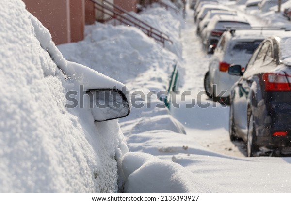 row of snow drifted cars in a row along the street\
near residential building at winter day light snowfall, close-up\
with selective focus