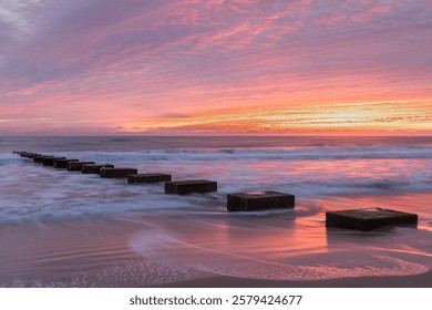 
A row of square concrete blocks extend into the ocean, leading toward a vibrant sunrise painting the sky pink and orange.  Gentle waves create soft patterns on a sandy beach on the Outer Banks NC.
