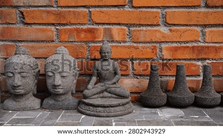 Row of Stone Buddha Statues and Decorations Against Red Brick Wall Background
A collection of grey volcanic stone sculptures arranged on a tiled floor against a textured red brick wall. The display features two Buddha heads, a full seated meditating Buddha statue, and three cone shaped stone ornaments (stupikas). The rough texture of the stone contrasts beautifully with the warm tones of the bricks. Ideal for concepts related to Buddhism, spirituality, meditation, garden decoration, rustic interiors, or Asian cultural heritage.