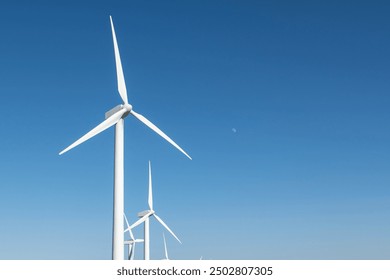 A row of tall wind turbines stand against a clear blue sky, with the sun shining brightly and a small moon visible. - Powered by Shutterstock