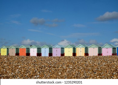 A row of ten colorful beach huts are in a line in the centre of the image in the foreground is a pebble beach above is a big blue sky with white clouds the sun is shining, Brighton, Sussex, UK.