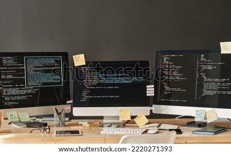 Row of three computer monitors with coded data and notepapers on their screens standing on table in office of IT support managers