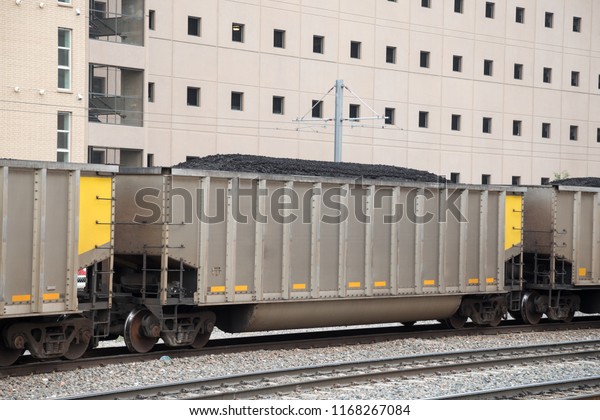 Row of train cars transporting coal, with an\
industrial building in the\
background