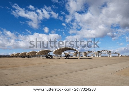 Row of twin engine multirole fighter jets parked under arched sun shelters on a concrete airbase apron under partly cloudy sky