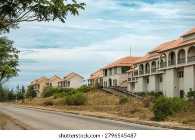 A row of vacant multi-story structures with arched balconies sits on a grassy hill next to an empty asphalt street under a cloudy sky.