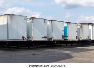 A row of white transport trailers are lined up outside a warehouse on a partly cloudy, sunny day.