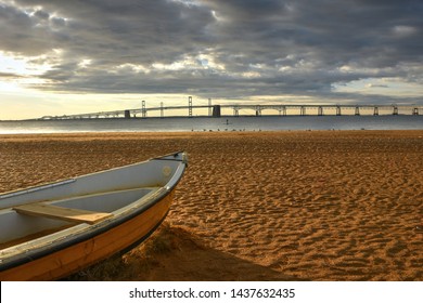 A Rowboat Moored On A Sandy Beach Points Toward The Expansive Chesapeake Bay Bridge.  A Scenic, Early Morning Landscape Along An Empty, Tranquil Shores Of The Bay On A Cloudy, Blue Sky Summer Day.