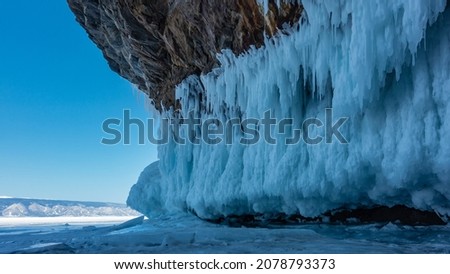 Rows of bizarre icicles hang from the base of the granite rock. Fragments of ice are scattered on the surface of the frozen lake. A mountain range against the blue sky in the distance. Baikal.