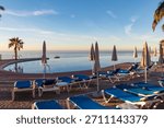 Rows of empty blue sunbeds sit facing a calm ocean beside a luxury swimming pool during early morning. This resort scene in Almunecar invites travelers to relax under the warm Andalusian sun.