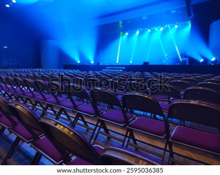 Rows of empty chairs face a stage illuminated by dramatic blue and white lighting. The setup suggests anticipation for an upcoming event or performance in a concert hall.