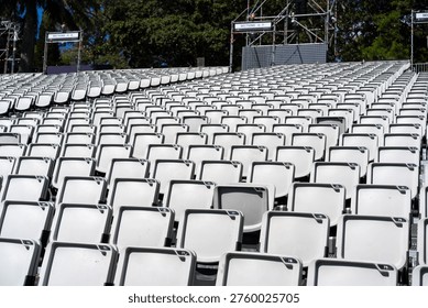 Rows of empty white numbered stadium seats stretch into the distance at an outdoor temporary event venue with metal scaffolding and green trees.