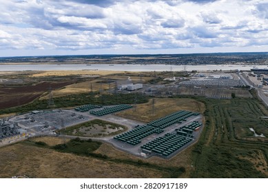 Rows of green battery containers and electrical power infrastructure stand on open land near a large body of water under a cloudy sky during the bright daylight hours in rural England
