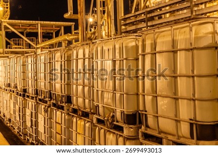 Rows of Intermediate Bulk Containers (IBCs) stacked in an industrial chemical plant at night. These large tanks are used for storing and transporting liquids or bulk materials in a manufacturing facil