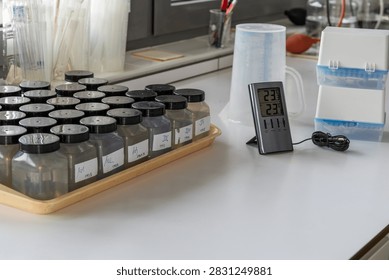 Rows of labeled sample bottles containing water on tray in laboratory. digital indoor-outdoor thermometer. The scene reflects environmental testing, quality control, and scientific sample analysis.