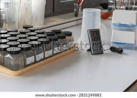 Rows of labeled sample bottles containing water on tray in laboratory. digital indoor-outdoor thermometer. The scene reflects environmental testing, quality control, and scientific sample analysis.