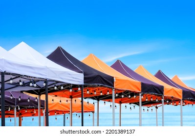 Rows of many foldable canopy tents with hanging LED light bulbs decoration for outdoors event on the beach against sea view background