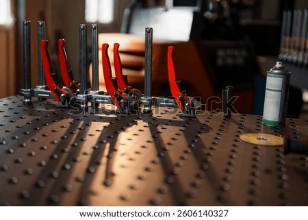 Rows of red toggle clamps resting on a metal welding table with holes, creating intricate shadows in the warm, inviting light of a busy workshop environment