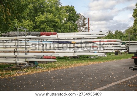 Rows of rowing boats (racing shells) and sculls stacked on outdoor metal racks at a riverside boatyard in Molesey, Surrey, England